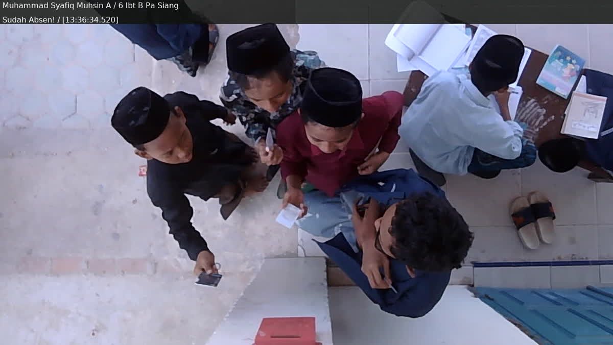 Rekaman kamera titik absensi: murid duduk di meja dengan buku, nama murid dan keterangan sudah absen muncul di layar kamera.